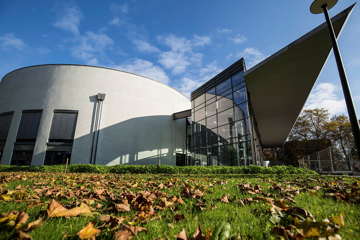 Modernes Architekturbauwerk mit weit vorspringendem halbrunden Glas-/Stahlbau, Grünfläche und Herbstlaub im Vordergrund.